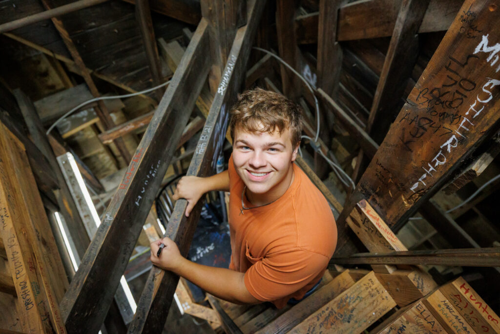 A student in an orange T-shirt smiles while writing his name on a wooden beam in the interior of the Administration Building.