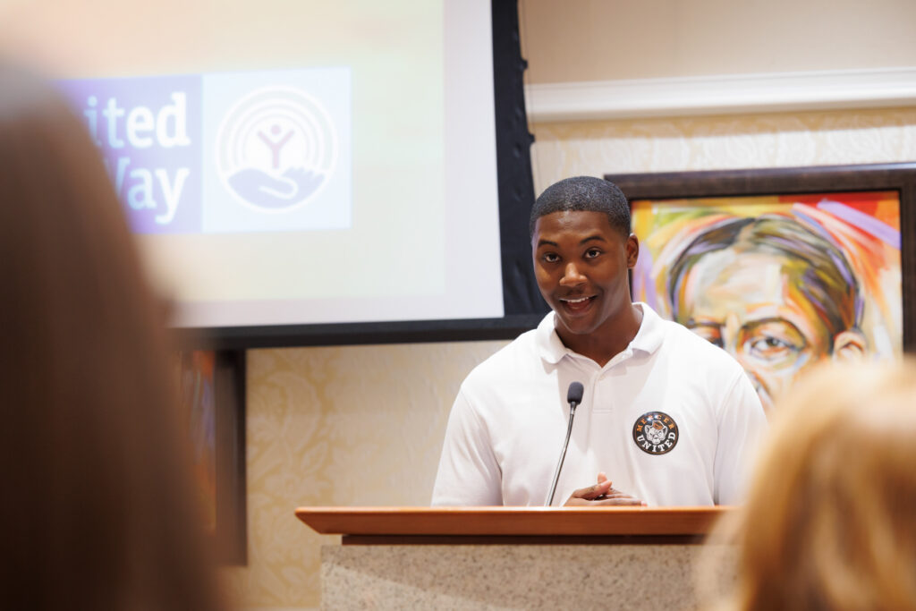 A man in a white polo shirt speaks at a podium in front of a United Way logo.