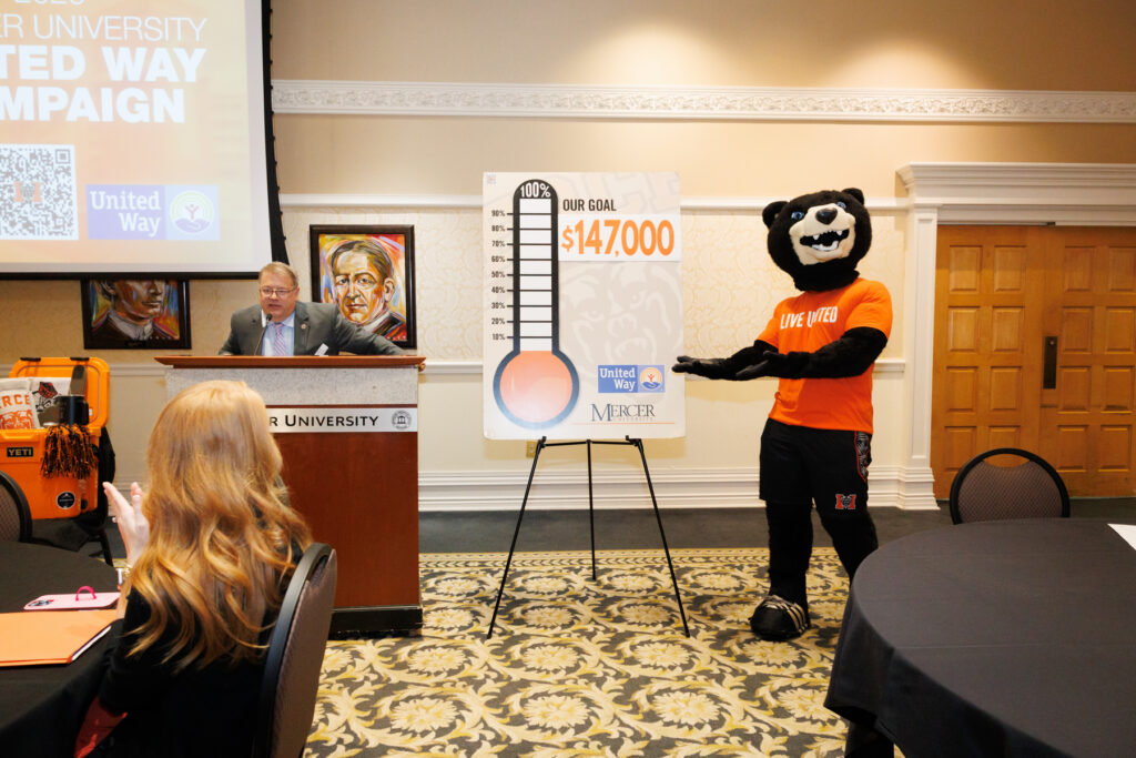 A man speaks at a podium next to Toby pointing to a fundraising thermometer showing $147,000.