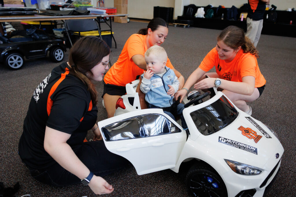 Two volunteers in orange shirts help a toddler into a white toy car.