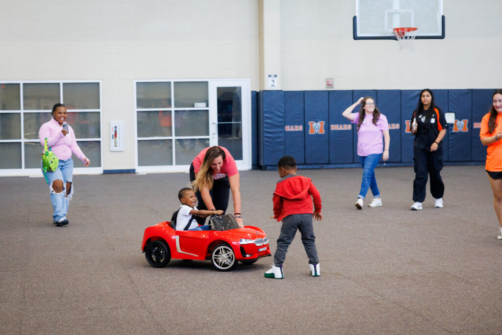 A young boy sits in a red toy car guided by a woman, with other people standing in a gymnasium.