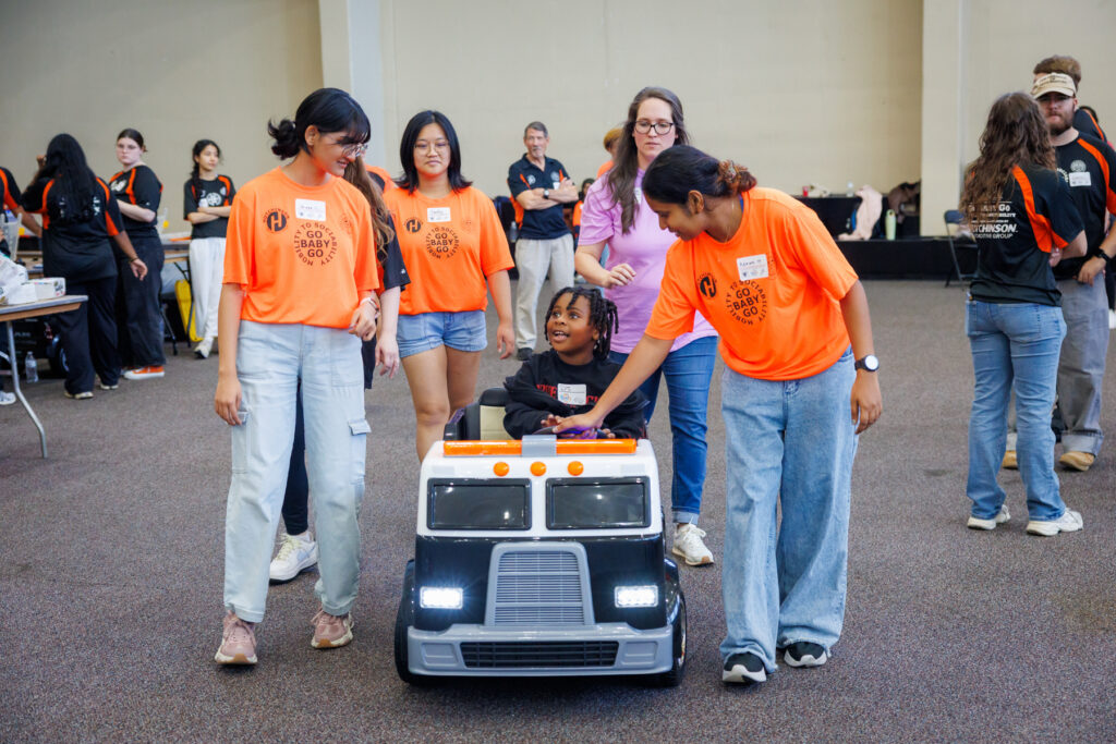 A child sits in a small toy truck while two volunteers in orange shirts guide them across a room.