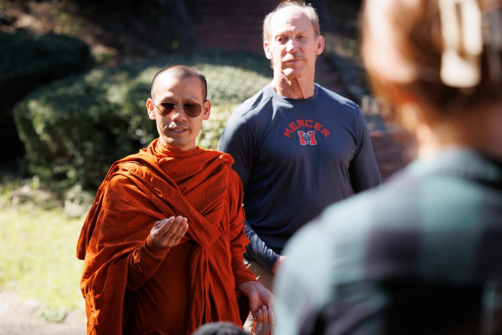 A monk in saffron robes speaks to a crowd, with a man in a Mercer University shirt beside him.