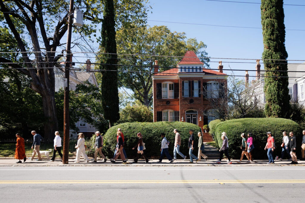 A diverse group of people walk in a line along a sidewalk past a brick house.