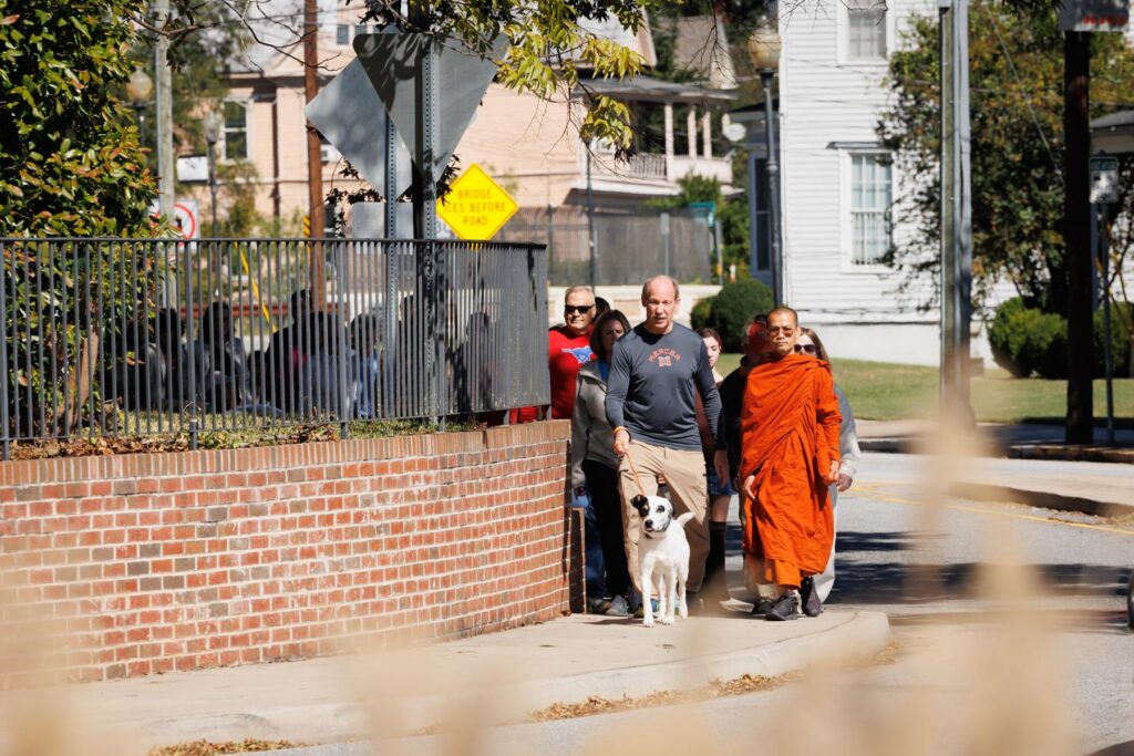 A person in orange robes walks with a group of people and a dog on a sidewalk.