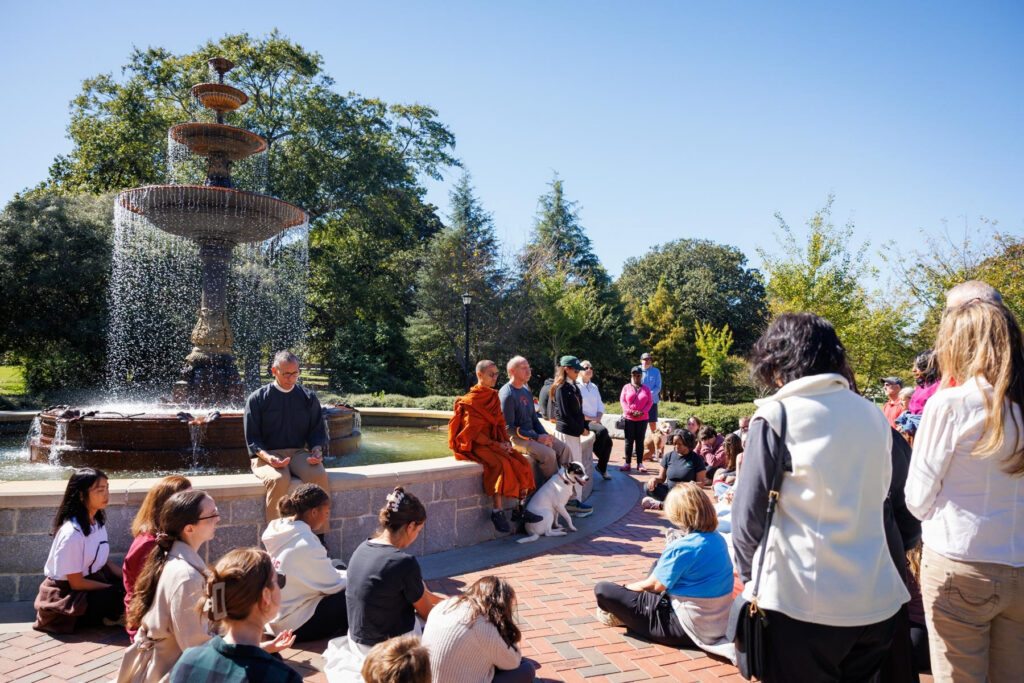 A group of people sit on the ground and a stone wall near a fountain, listening to a monk.