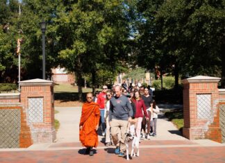 Monk teaches meditation, leads Community Peace Walk during Mercer visit A group of people walking through a brick campus gate, including a monk in orange robes and a man walking a dog.