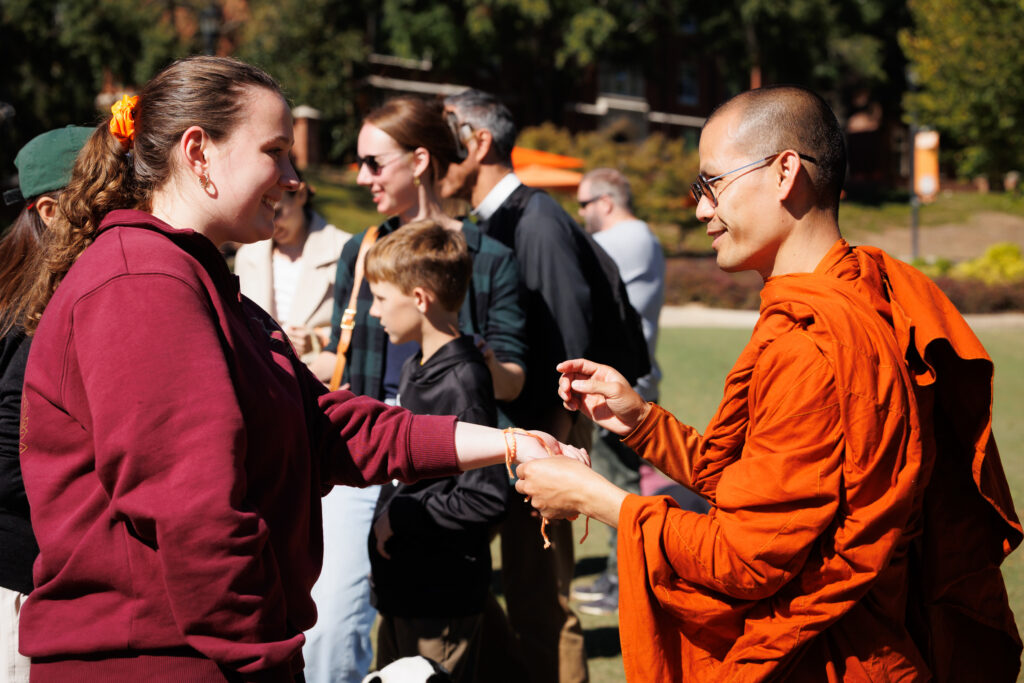 A monk in orange robes ties a string bracelet onto the wrist of a young woman.