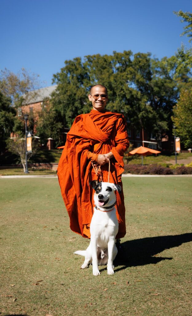 A Buddhist monk in saffron robes standing on a lawn, holding the leash of a white and black dog.