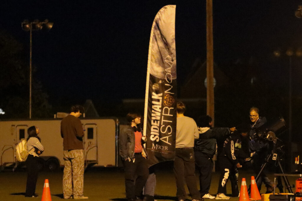 People gathered at night around a Sidewalk Astronomy banner and a telescope.