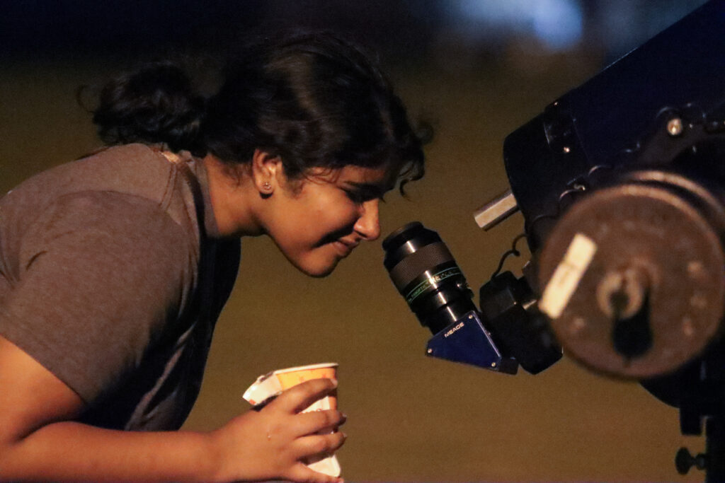 A person looking through the eyepiece of a telescope while holding a paper cup.