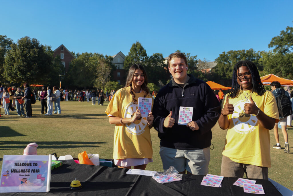 Three people stand behind a wellness fair sign and table, holding Wellness Bingo cards.