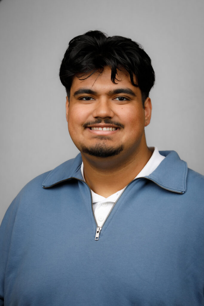 A smiling man with dark hair and a goatee wearing a blue quarter-zip sweater.
