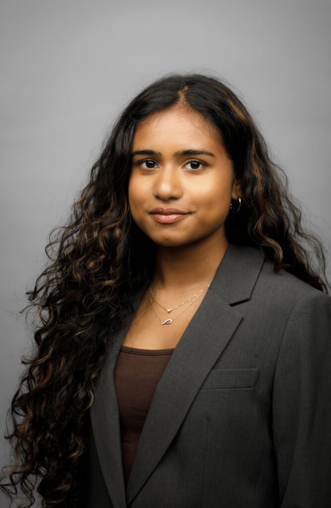 A young woman with long, dark wavy hair wearing a grey blazer.