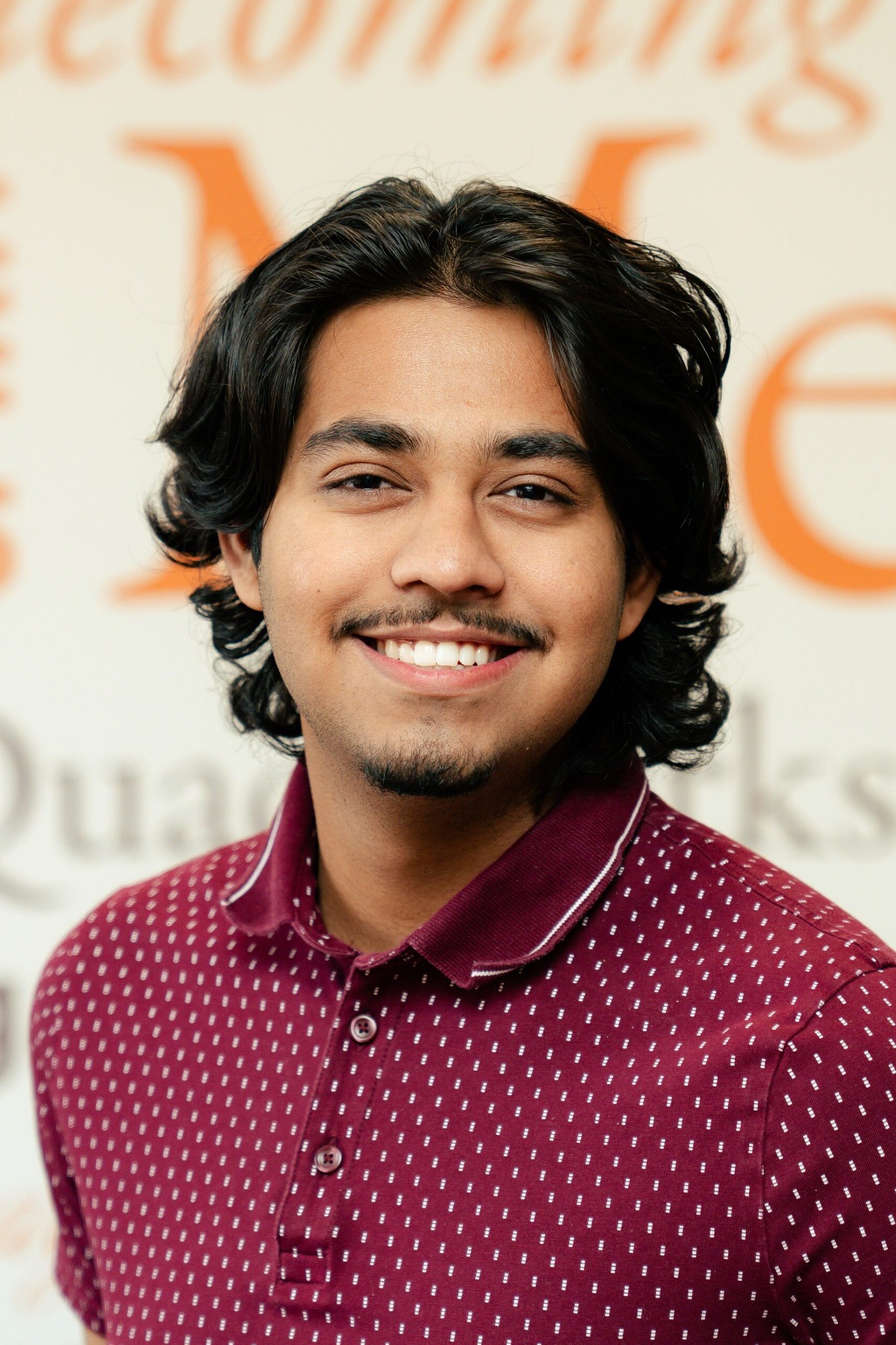 A young person with shoulder-length dark hair and a mustache smiles in front of a branded wall.