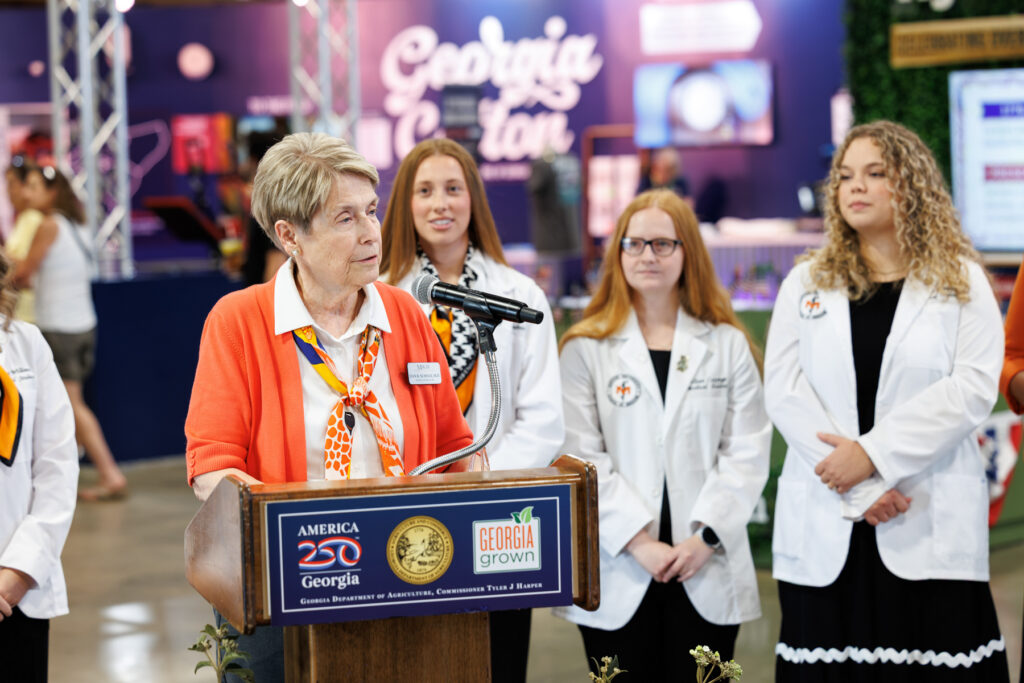 School of Medicine Dean Jean Sumner speaks at a podium with three students in white medical coats standing behind her.