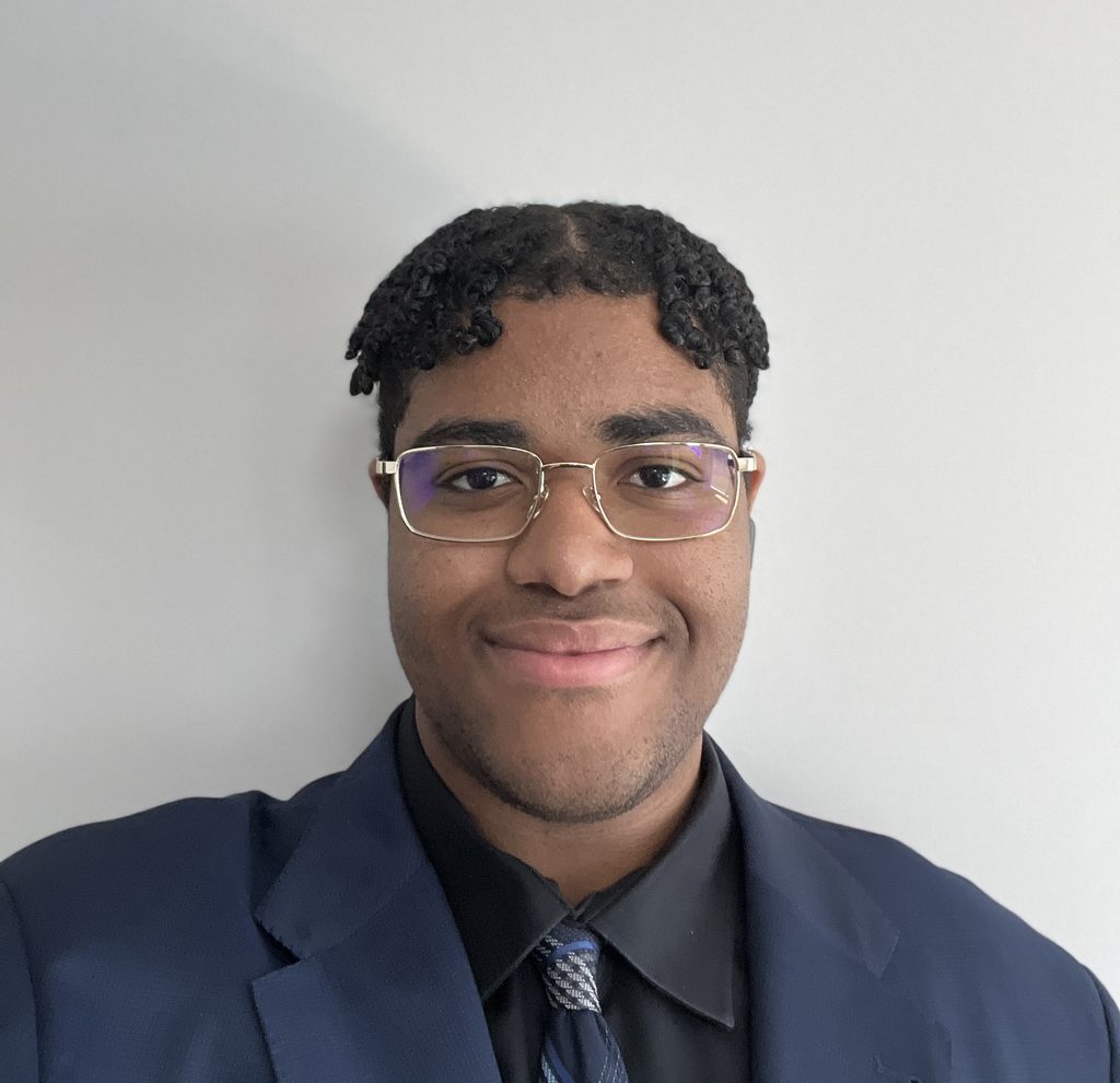A young man wearing glasses, a navy blue blazer, a black shirt, and a patterned tie.