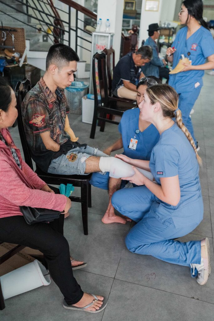 A medical worker in scrubs fits a prosthetic leg on a seated man with amputated limbs in a clinic setting.