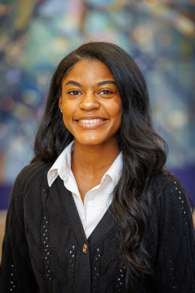 A young woman with long dark hair smiles at the camera, wearing a white collared shirt under a black cardigan.