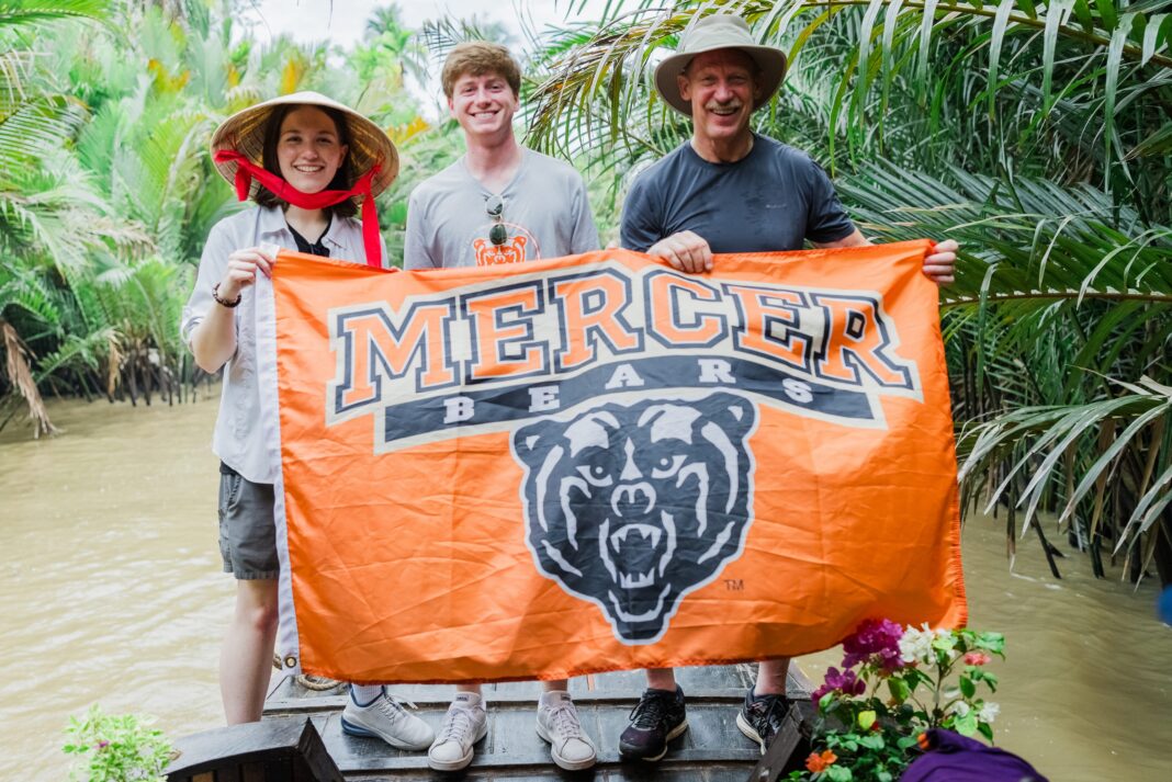 MOMVietnam2024-208 Three people stand on a wooden boat holding an orange Mercer Bears flag in a tropical riverside setting.
