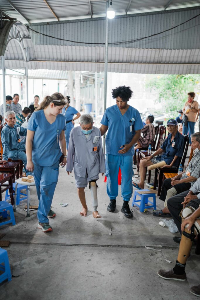 Two healthcare workers assist a patient with a prosthetic leg as others sit and observe in a clinic setting.