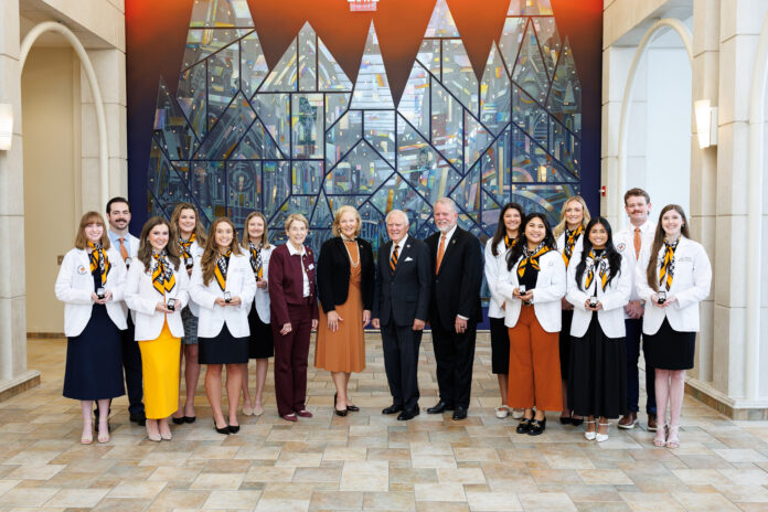 A group of students and faculty members in professional attire posing for a photo in front of a stained glass mural.