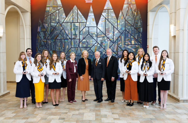 A group of students and faculty members in professional attire posing for a photo in front of a stained glass mural.