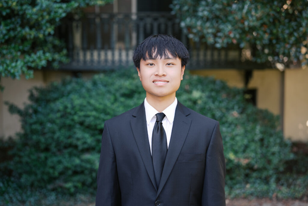 A young man in a black suit and tie standing outdoors.