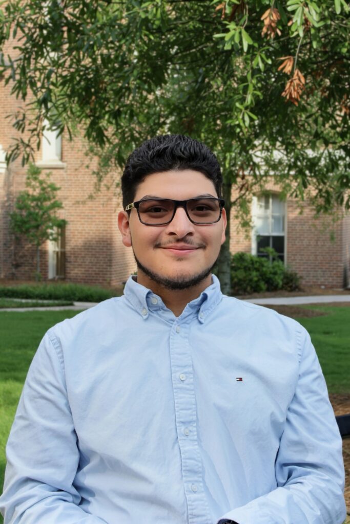 A young man with glasses and a light blue button-down shirt standing outdoors.