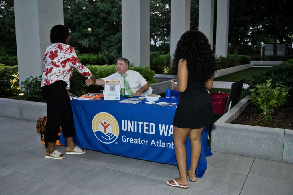 A man behind a United Way Greater Atlanta table shakes hands with a woman, while another woman stands nearby.