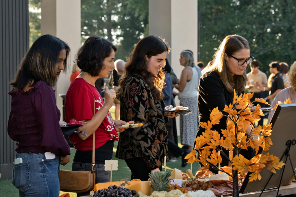 Four women stand at an outdoor buffet table selecting food.