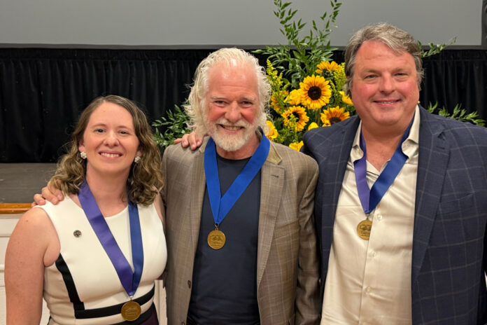 Three people wearing blue ribbons with gold medals pose for a photo in front of a sunflower arrangement.