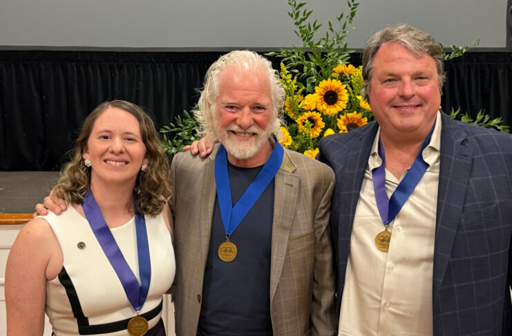 Three people wearing blue ribbons with gold medals pose for a photo in front of a sunflower arrangement.