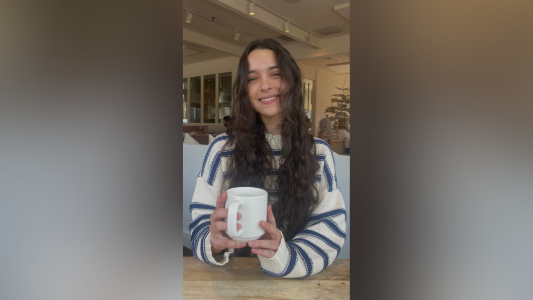 A woman with long wavy hair wearing a striped sweater smiles while holding a white mug at a cafe table.