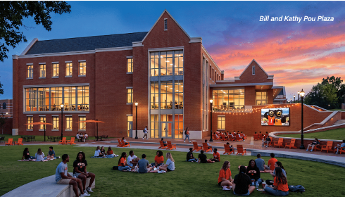 Students gather on a lawn at Bill and Kathy Pou Plaza in front of a brick building at sunset.