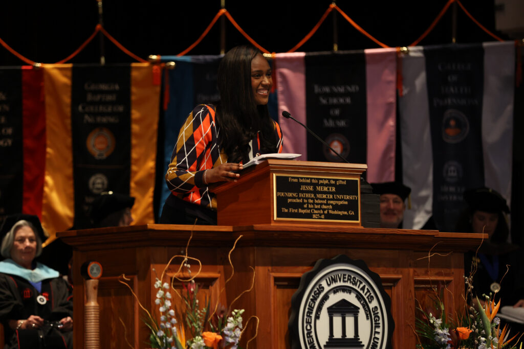 A woman speaks from a wooden podium at a formal ceremony, with academic banners in the background.