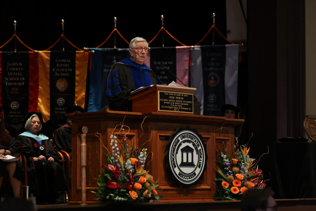 Kirby Godsey in academic regalia speaks from a wooden podium decorated with floral arrangements.
