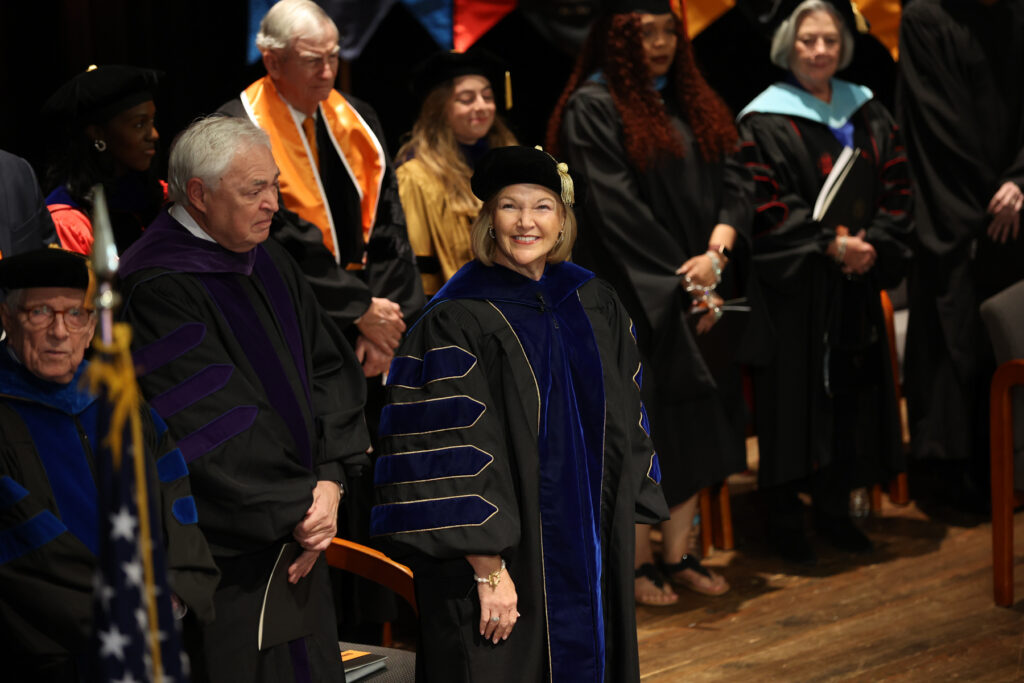 President Elkins academic regalia stands in the foreground, with other faculty members and graduates behind her on stage.