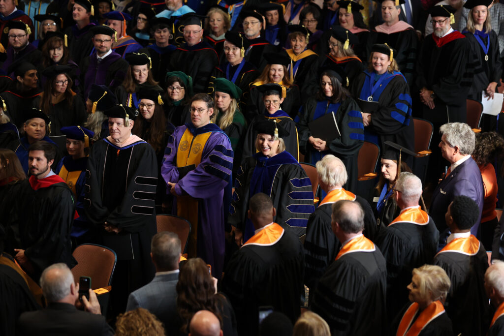 Faculty in academic regalia standing in a large auditorium with President Elkins in the center.