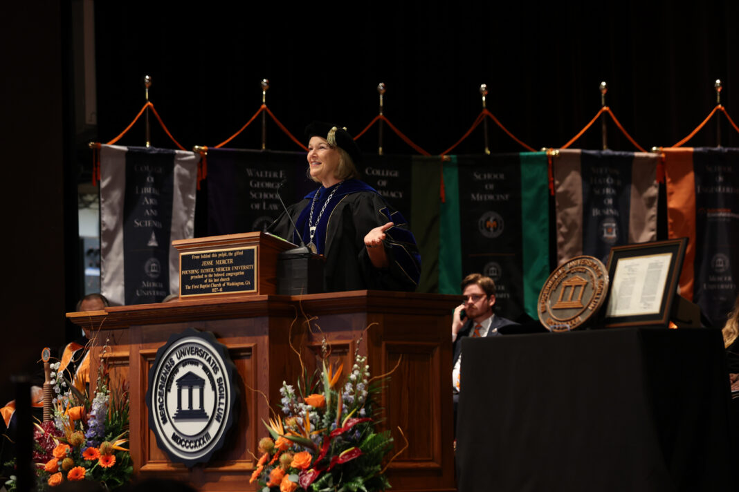 investiture for social-4 President Elkins in academic regalia speaks from a wooden podium at a university ceremony.