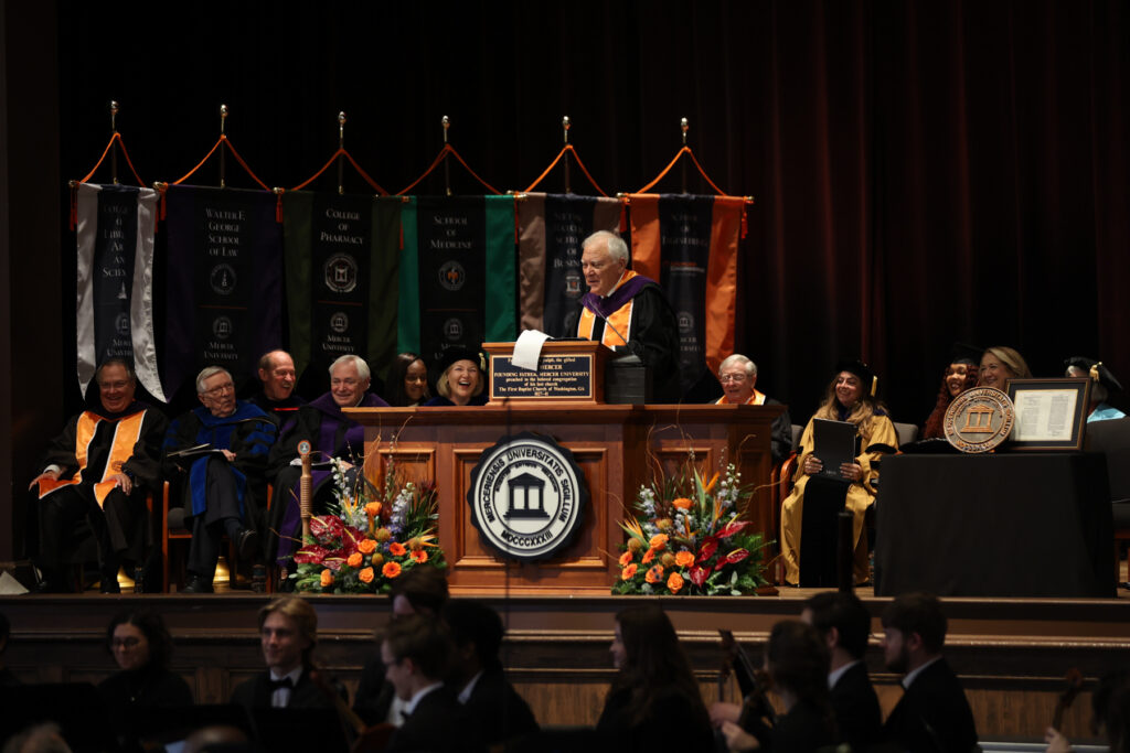 Former Georgia Gov. Nathan Deal in academic regalia speaks from a podium on a stage before a seated audience.