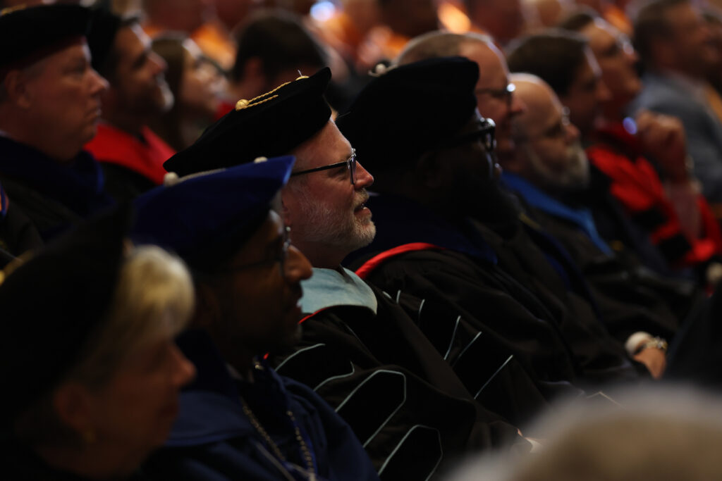 People in academic regalia sitting in a crowd during a ceremony.