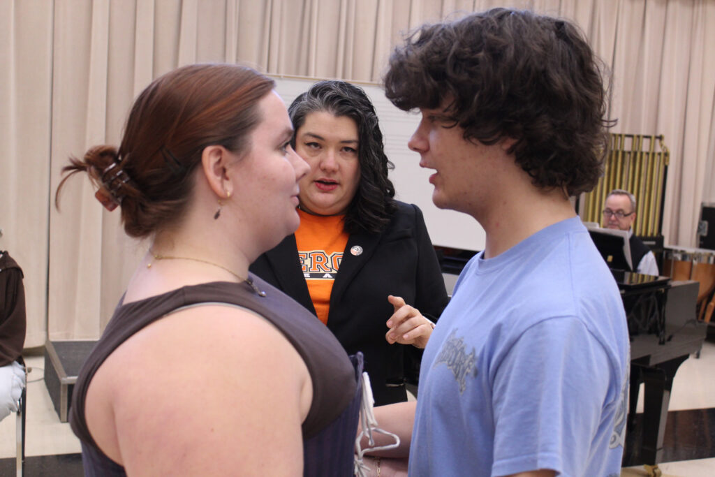 A woman and a young man in profile face each other as a woman in an orange shirt speaks to them.
