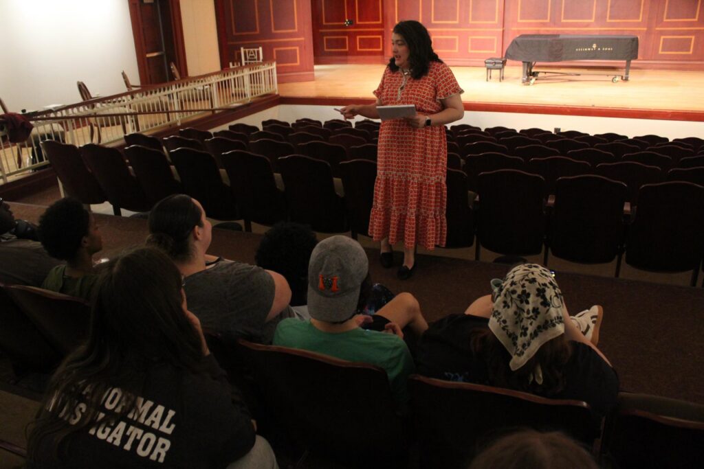 A woman in a patterned dress stands on a stage and addresses a seated audience in a theater.