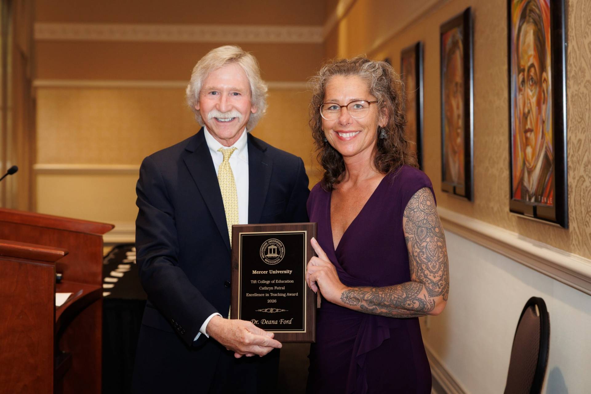 A man presents an award plaque to a smiling woman in a formal indoor setting with framed artwork on the wall.