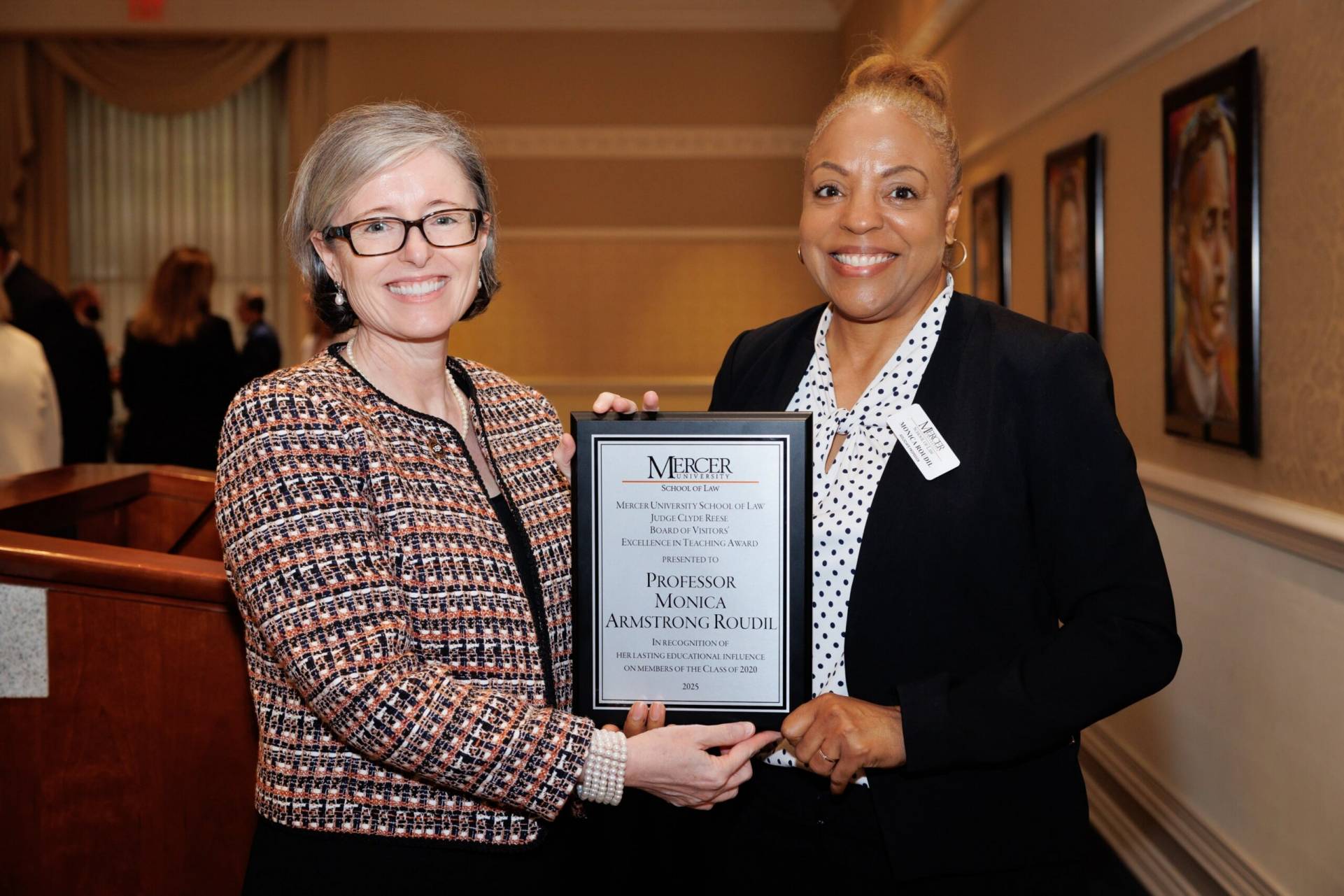 Two women stand indoors, one holding a plaque recognizing Professor Monica Armstrong-Roudil for excellence in teaching.