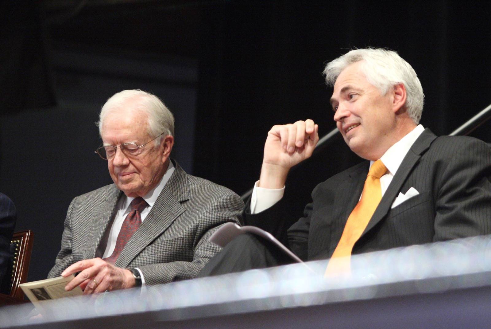Two men in suits sit side by side on a stage; one reads a book while the other looks ahead and smiles.