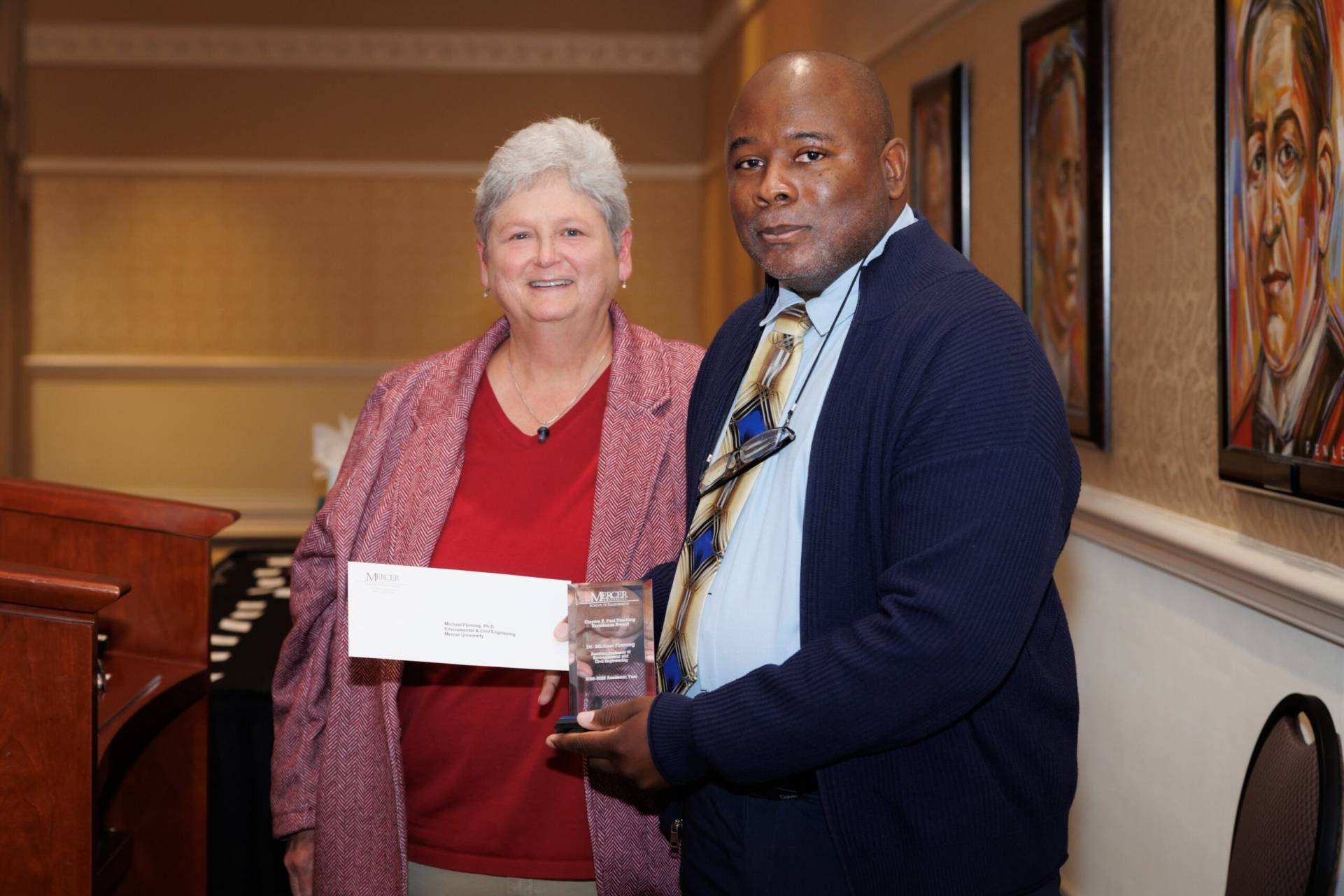 Two people stand indoors; one holds an envelope, and the other holds a glass award and wears a lanyard.