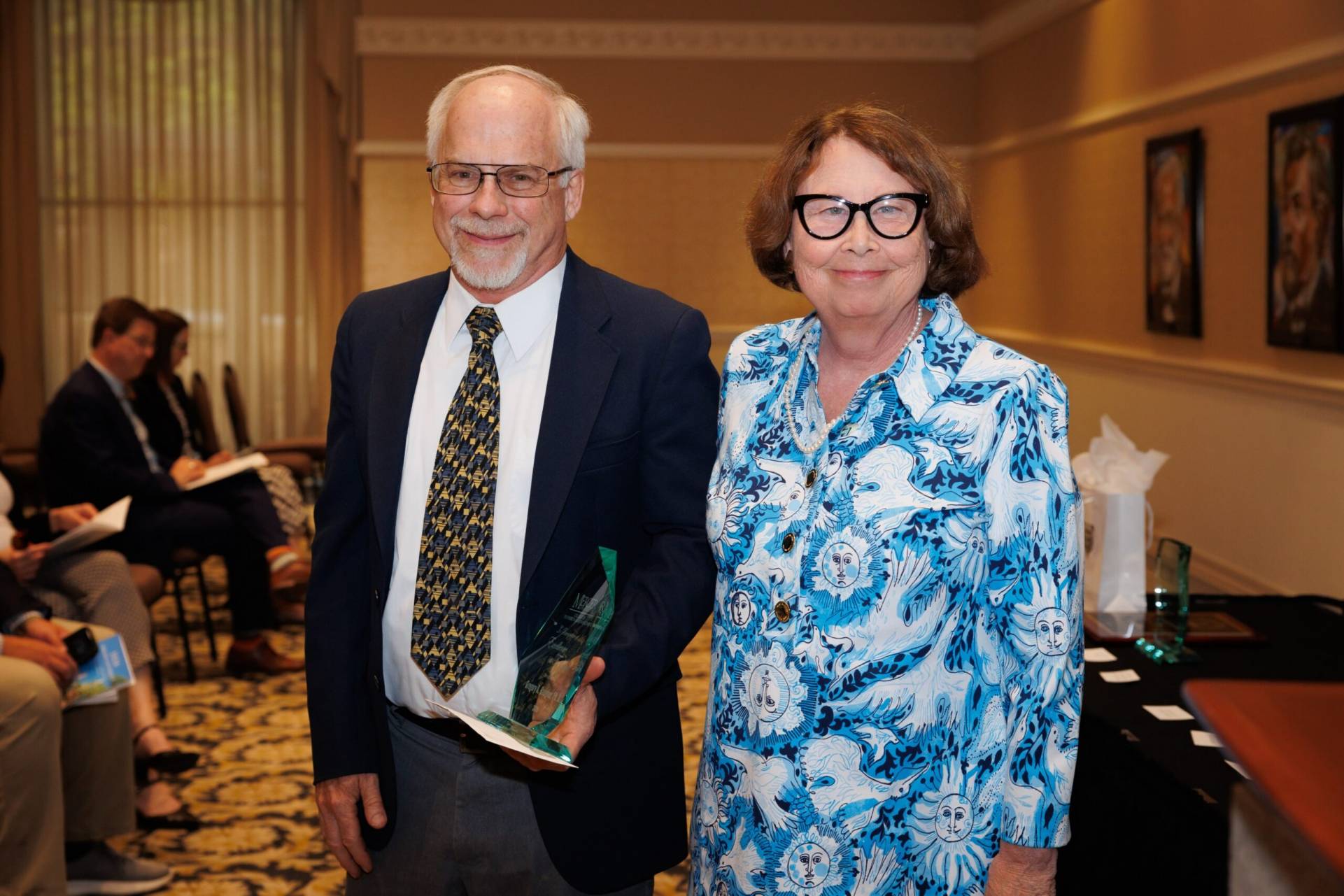 Two older adults stand indoors; one holds a glass award. People are seated in the background.