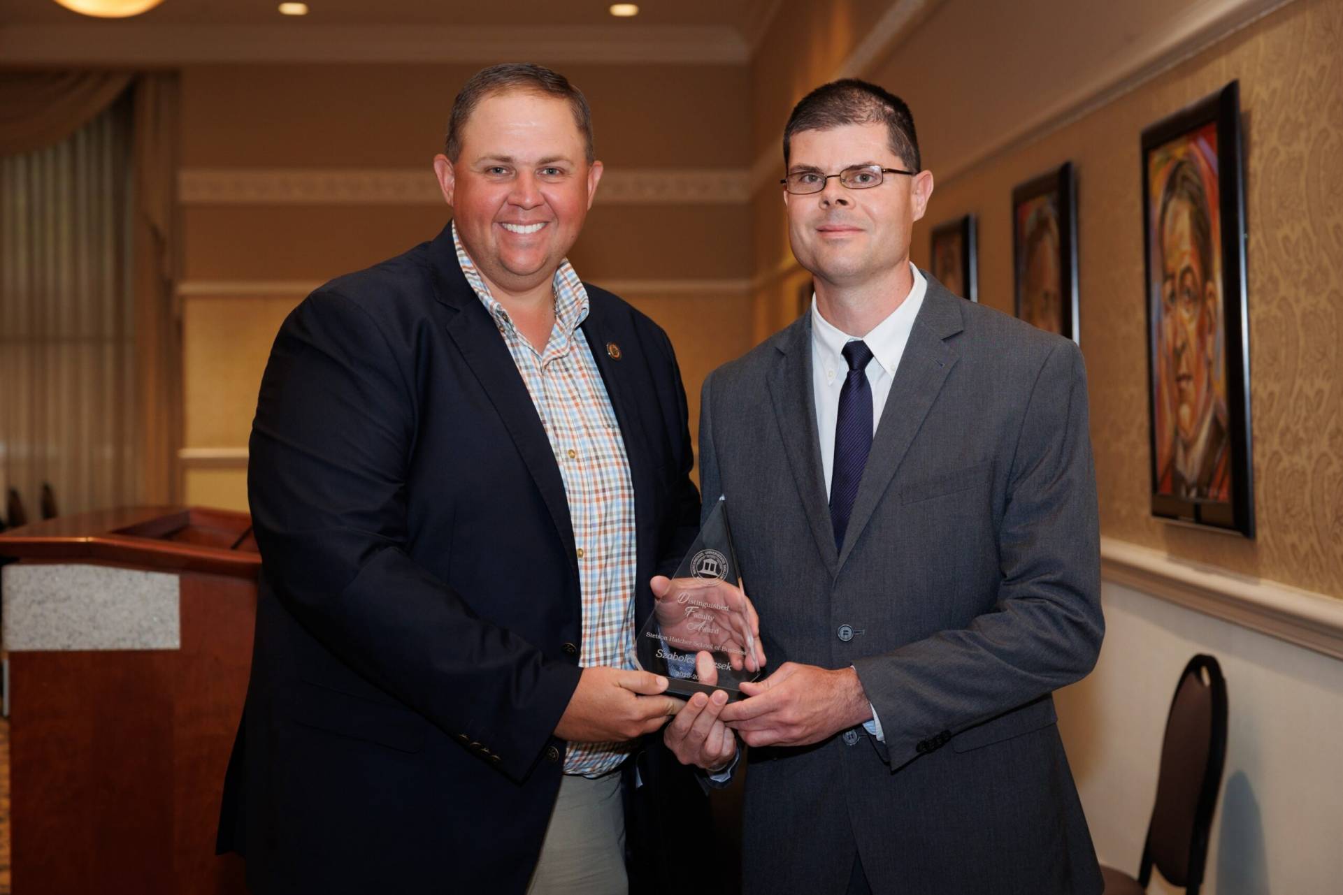 Two men in suits stand indoors, one presenting an award to the other, both facing the camera and smiling.
